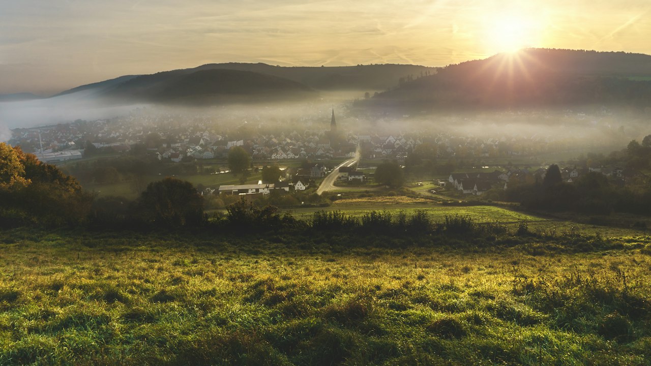 A hazy city skyline on green hills under a soft morning sky.